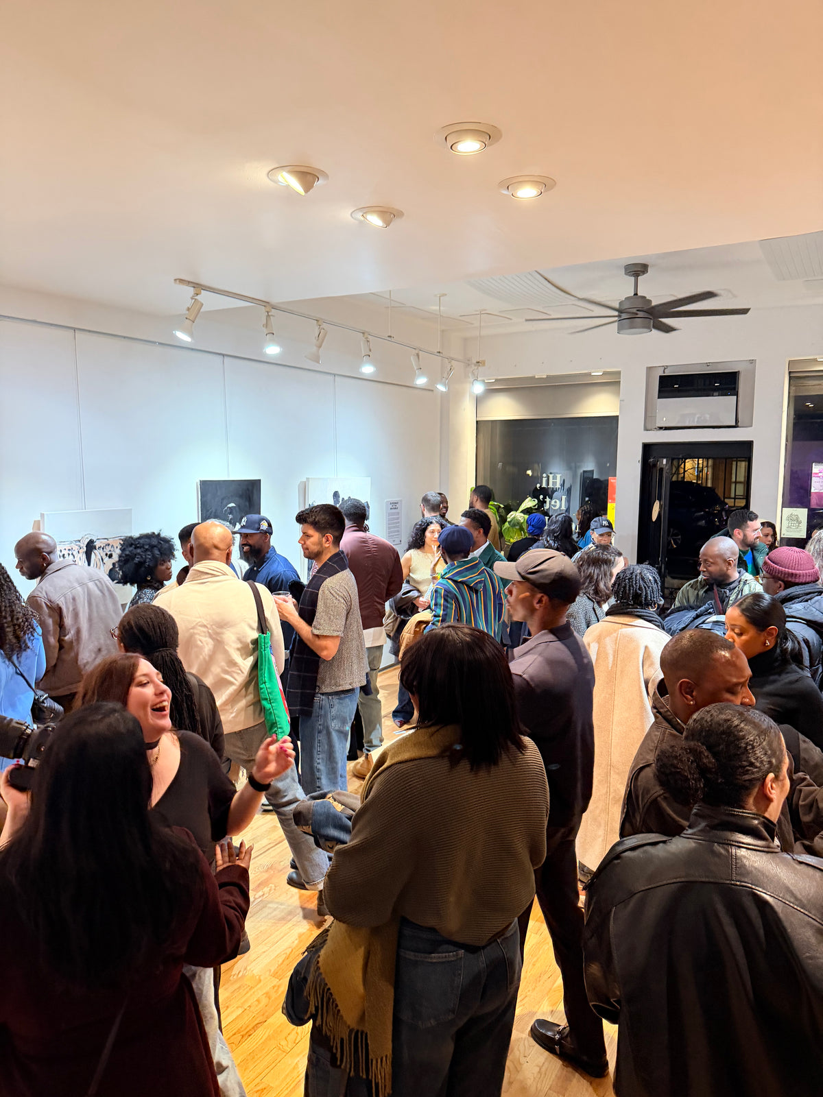 Dufie Kufuor's Art Exhibition. A crowded indoor gallery or studio space with many people standing and talking, viewed from above. The room has white walls, track lighting, a ceiling fan, and wood flooring.