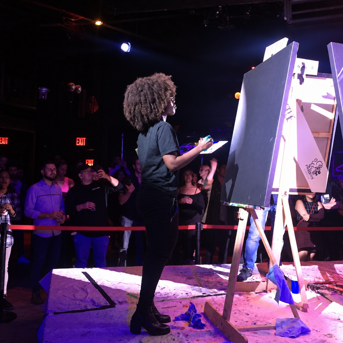 A woman with an afro stands at an easel painting on a canvas in a dimly lit event space, with a crowd of onlookers behind her.