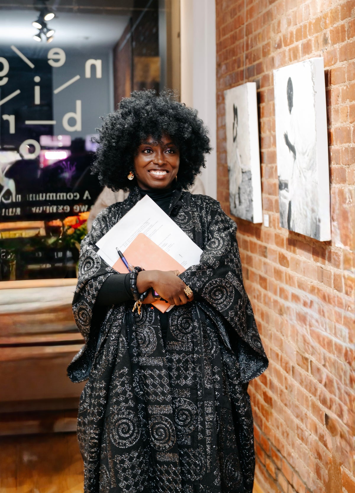 Visual Artist Dufie Kufuor smiling with a large black afro stands indoors against a brick wall, wearing a black patterned shawl or dress over a black top. She holds papers and a pen.